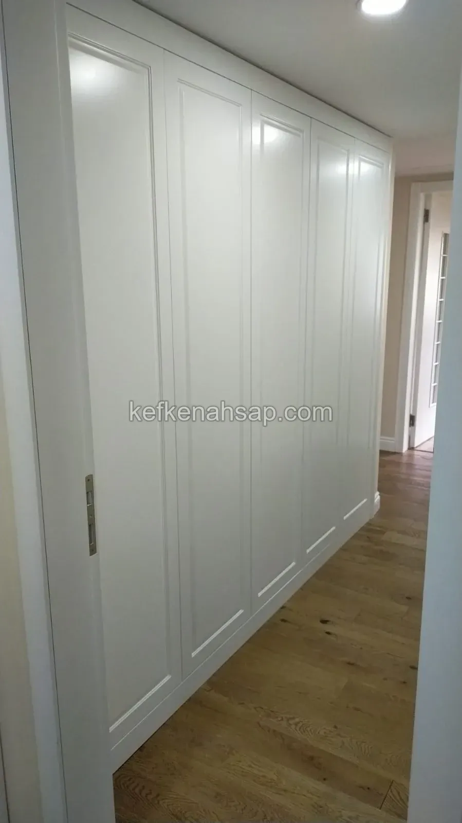 Koridor boyunca uzanan, çerçeveli kapaklı beyaz boyalı gömme dolap; zeminde ahşap parke var