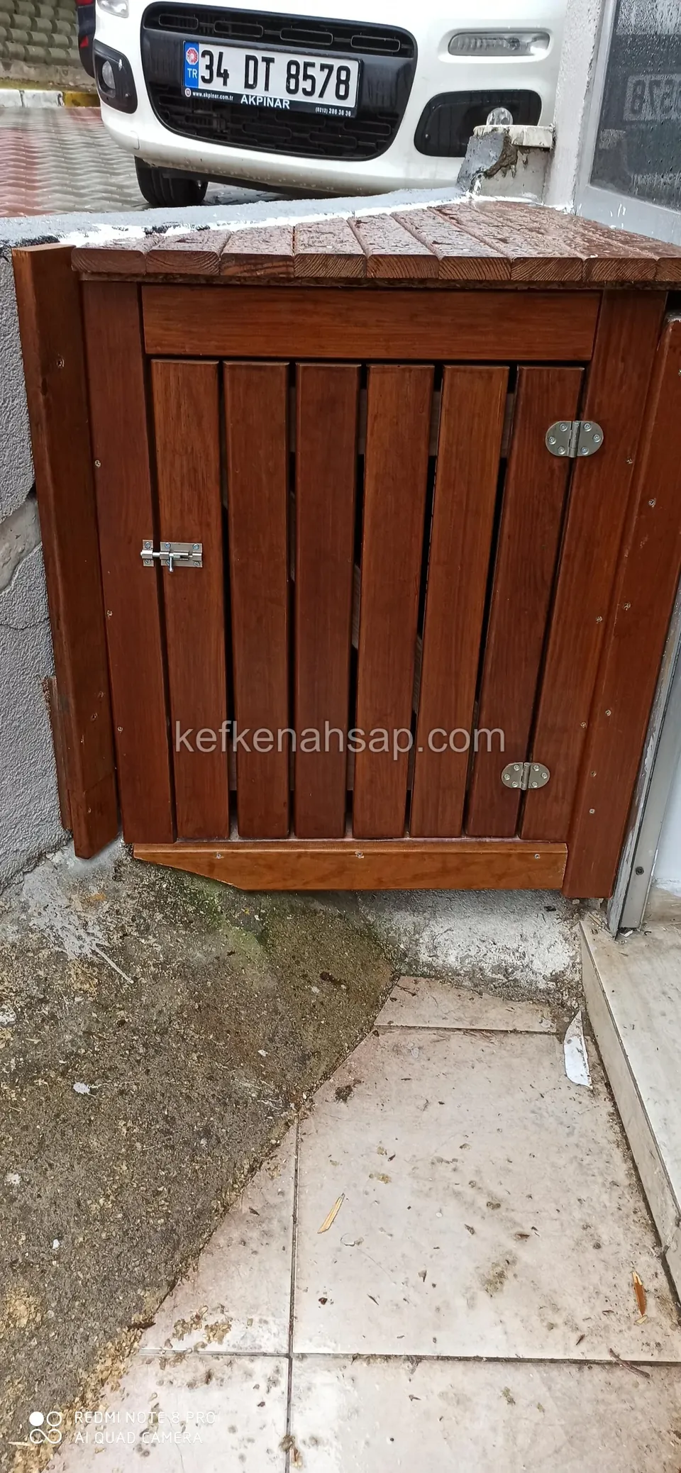 Eğimli kapaklı köşe dolabın dikey tahtalı kapağı, iki yanında metal sürgü ve menteşeler; kaldırım taşı zemin üzerinde
