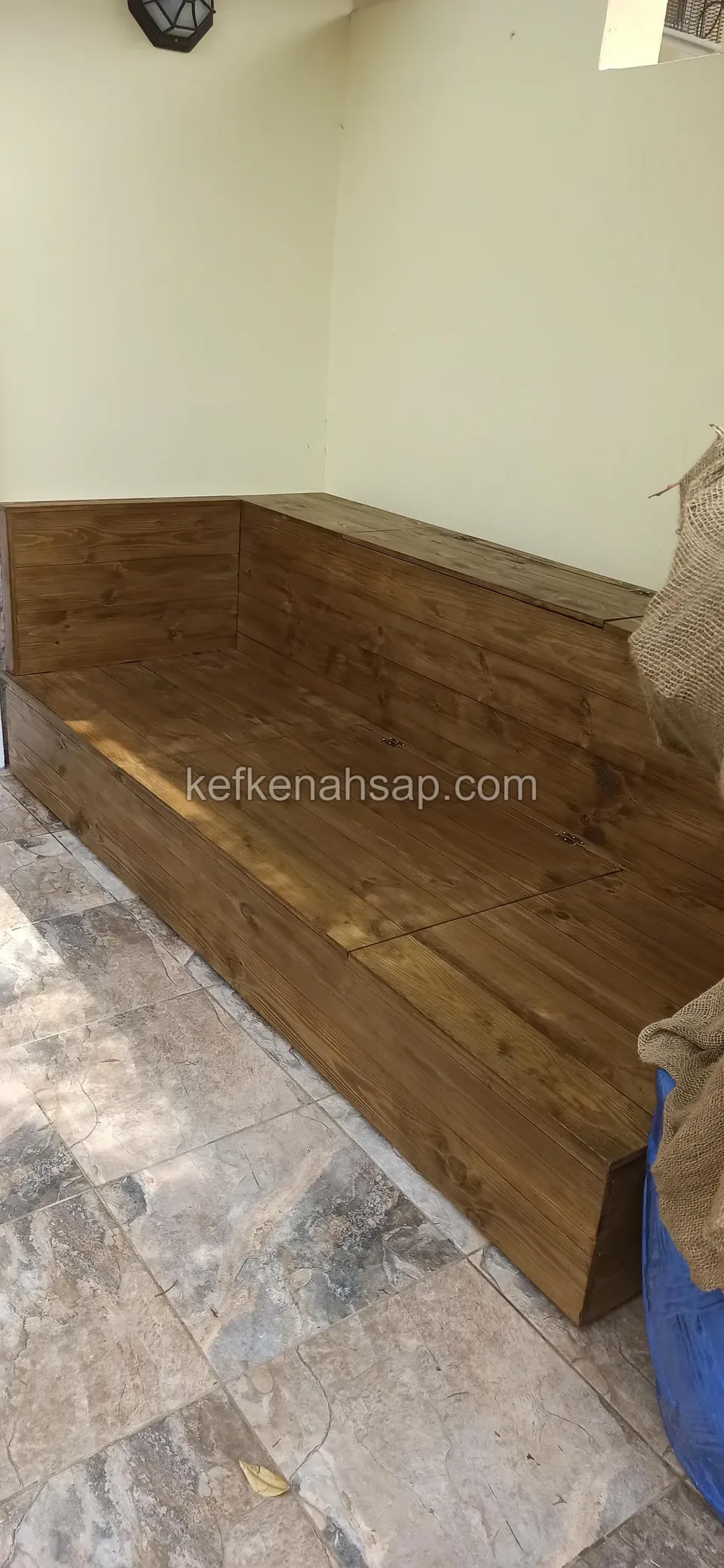 Eine Holzbank in der Ecke einer gelben Wand auf Steinplatten, von oben gesehen, daneben eine grüne Abdeckung