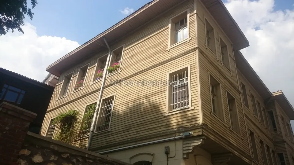 Alttan görünen, köşe yapan üç katlı bej boyalı ahşap kaplamalı bina, pencere parmaklıkları, geniş çatı saçağı