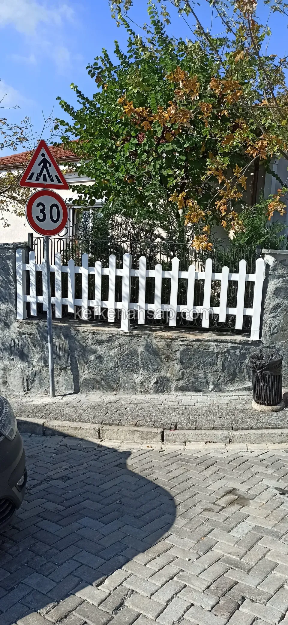 Köşe başındaki taş duvar üzerinde uzayan beyaz çıtalı çit, arkada sonbahar renkli ağaç ve levhalar