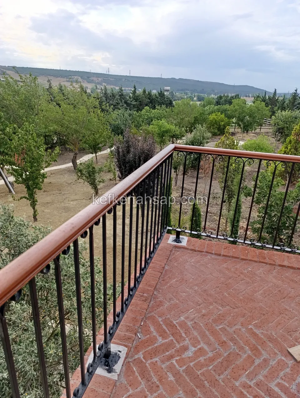 Black iron railing turning at a terrace corner with a brown timber handrail, trees and a valley behind