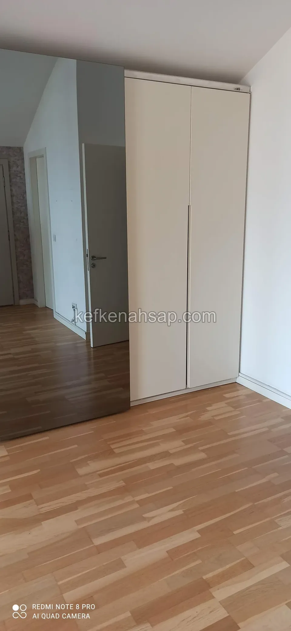 Odanın köşesinde tek parça görünen, krem renkli iki kanatlı yüksek dolap ve açık renk parke