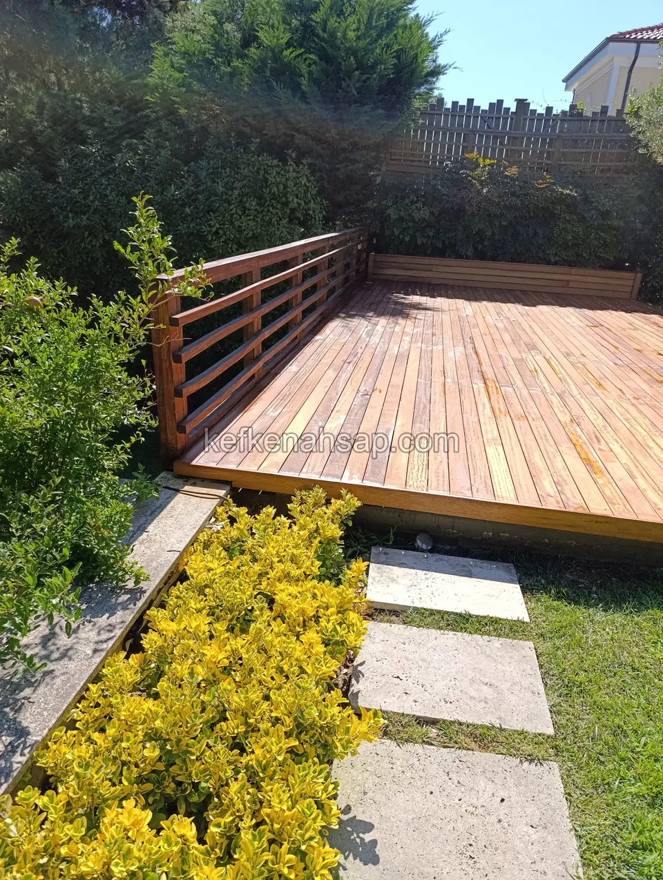 Orange-brown varnished deck with a horizontal-slat timber railing and a bench in the corner, with yellow shrubs