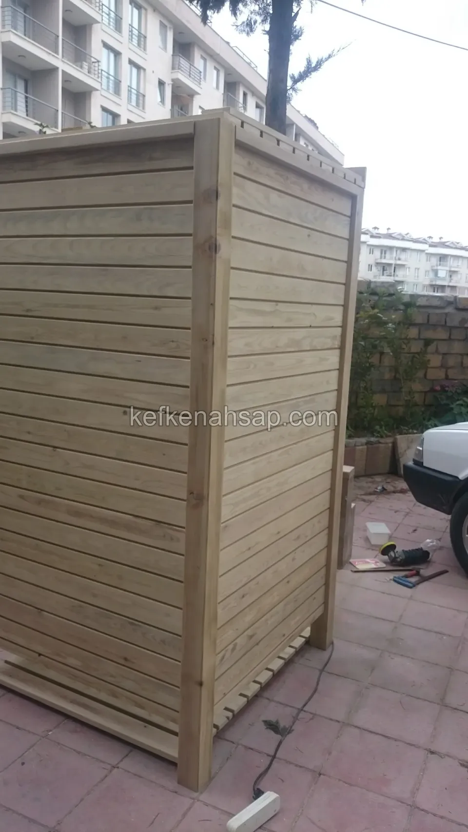 Bina önünde, köşeden görünen yüksek ahşap kasa; yan ve ön yüzü yatay tahtalı, yerde kablo var