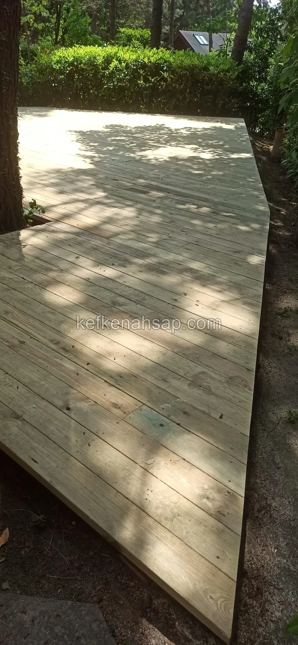 Çam ağacının yanında, açılı kenarlarla çakıllı zemine oturan geniş ve açık renkli deck