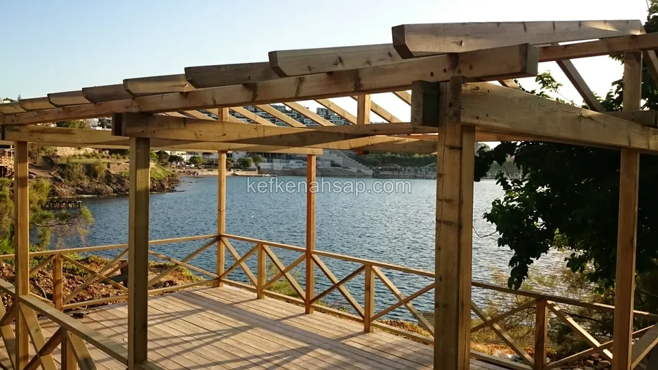Sakin bir koya bakan, çapraz korkuluklu ahşap deck ve üzerindeki kalın kirişli pergola