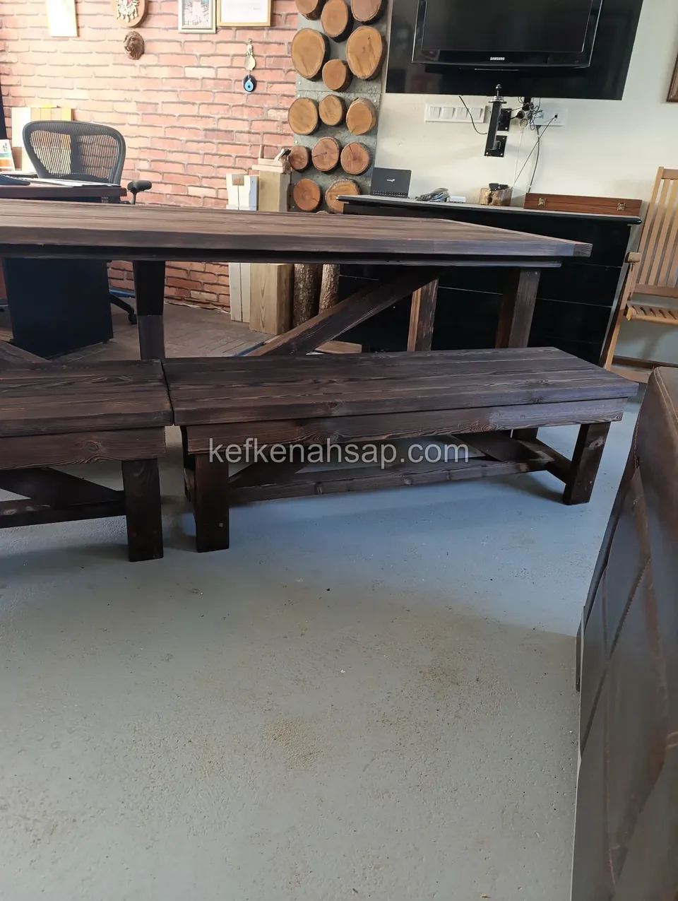 A dark-varnished timber table with two benches side by side in front of it, with a brick wall