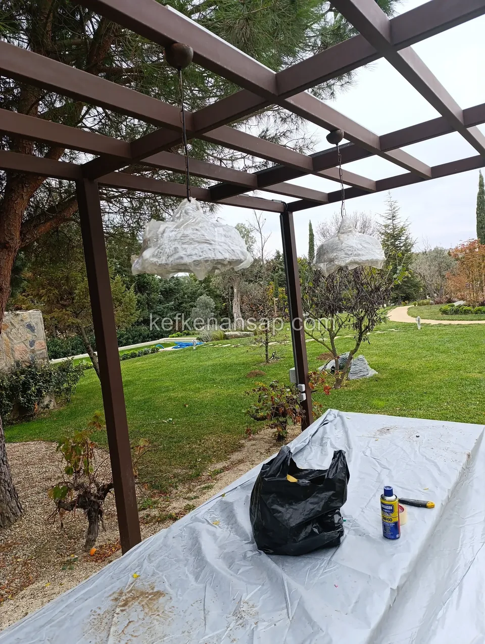 Bahçede koyu boyalı ahşap pergola kirişleri, altında serilmiş plastik örtü, sprey kutusu ve poşet