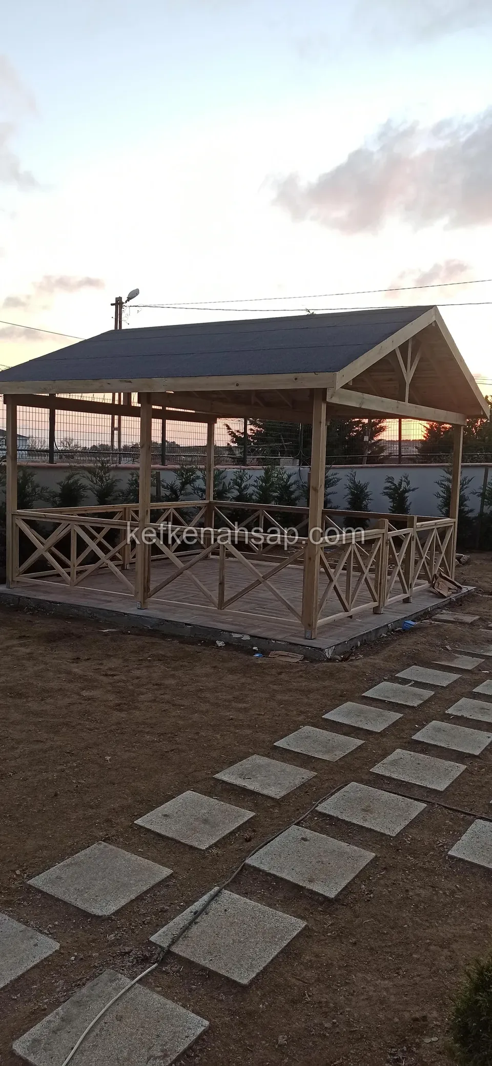 Koyu renk çatı kaplı korkuluklu kamelya, önünde sıralı beton basamak taşları ve düzenlenmemiş toprak