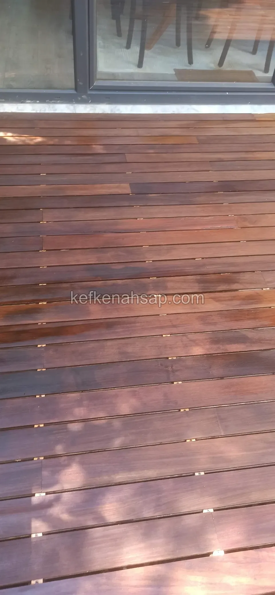 Dark red-brown deck boards in front of a glass door, showing even gaps and the joint lines at the board ends