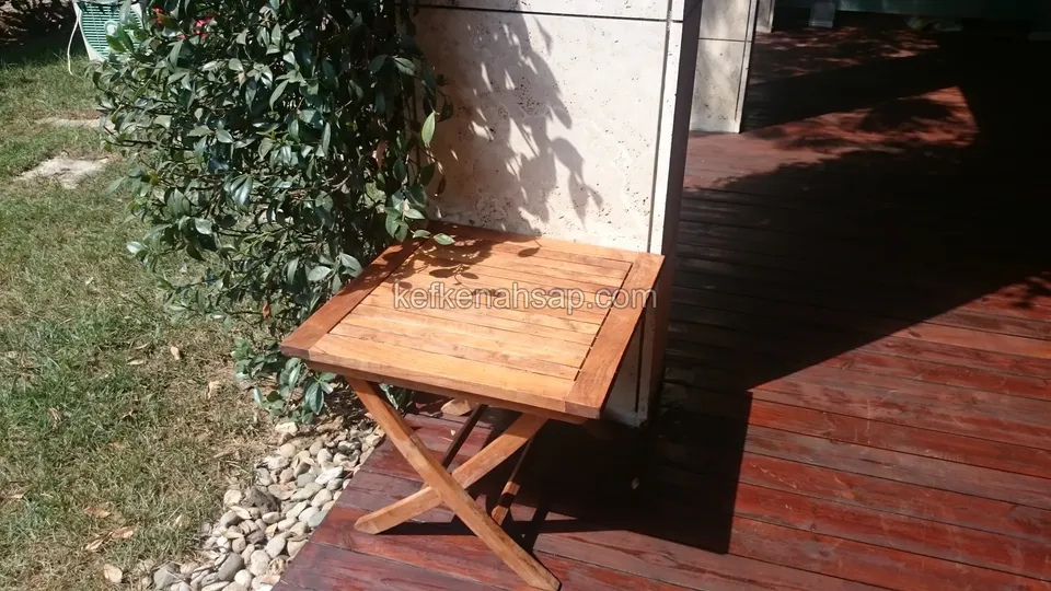 Zwischen dunklem Holzdeck und Rasen steht ein kleiner quadratischer Klapptisch aus Holz mit gekreuzten Beinen