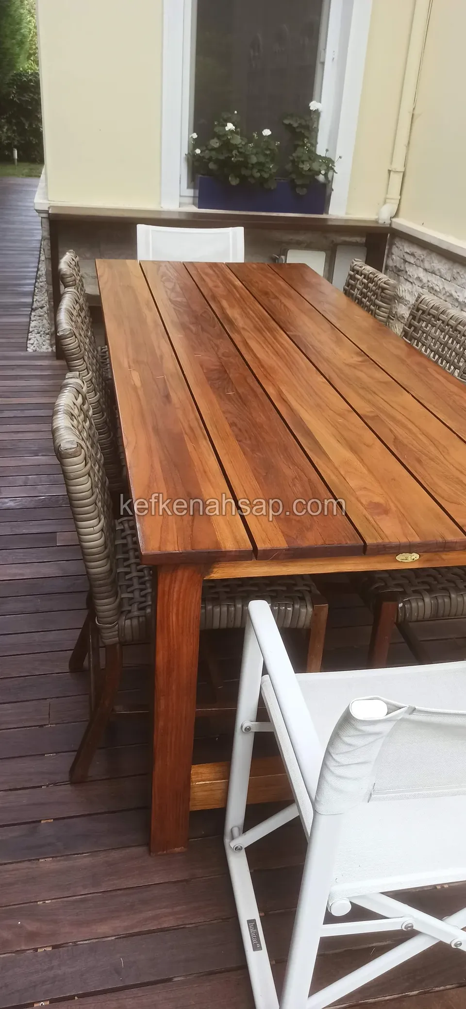 Koyu renkli deck terasta uzun ahşap yemek masası, çevresinde hasır ve beyaz sandalyeler