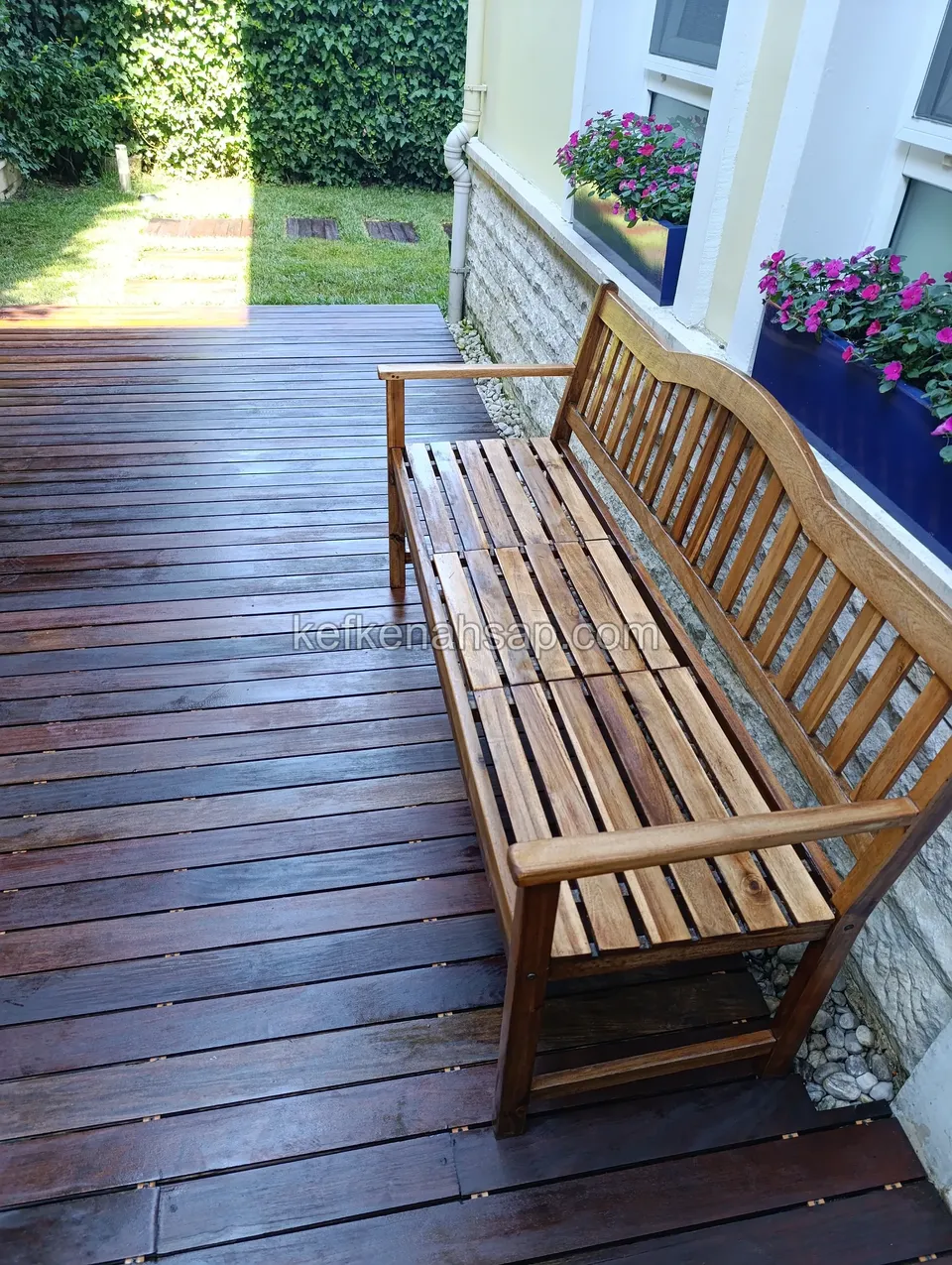 Eine lange Holzgartenbank mit geschwungener Lehne auf dunkelbraunem Deck entlang einer steinverkleideten Hauswand