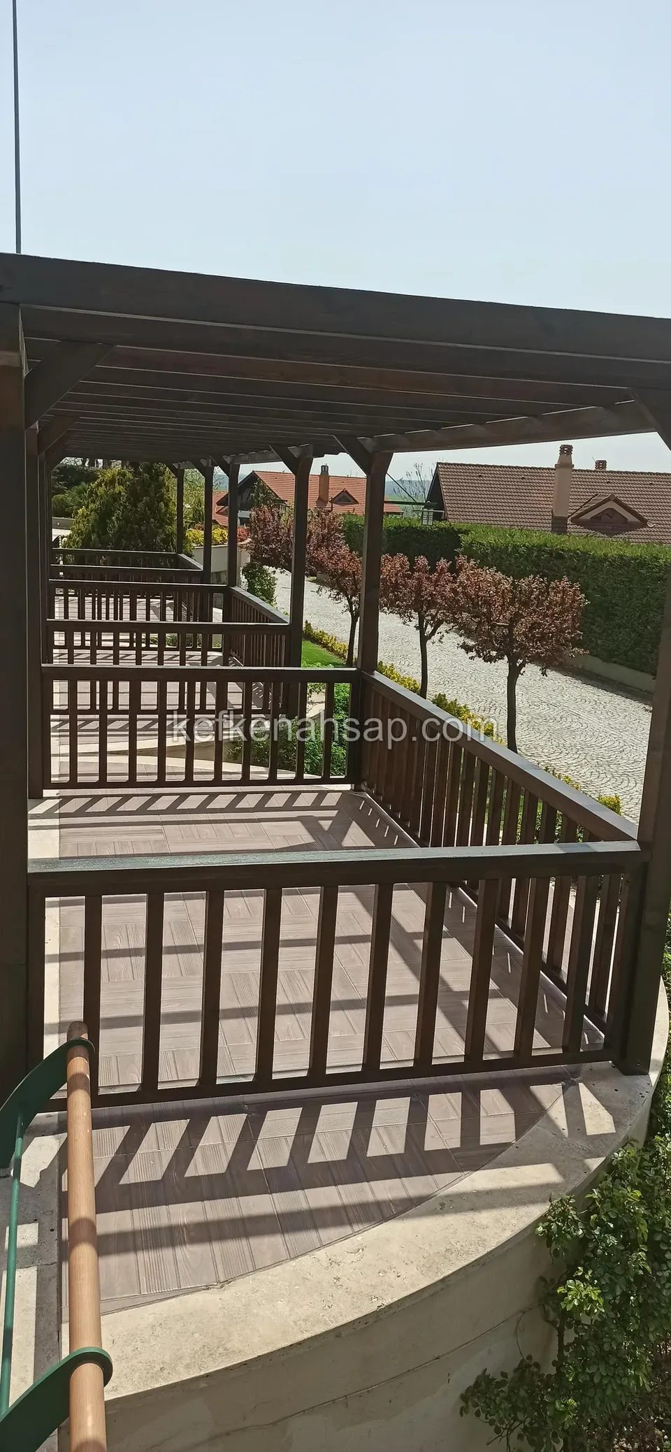 Koyu kahverengi ahşap pergola ve çıtalı korkuluk, yanında çakıllı yol ve fidanlar, uzakta evler