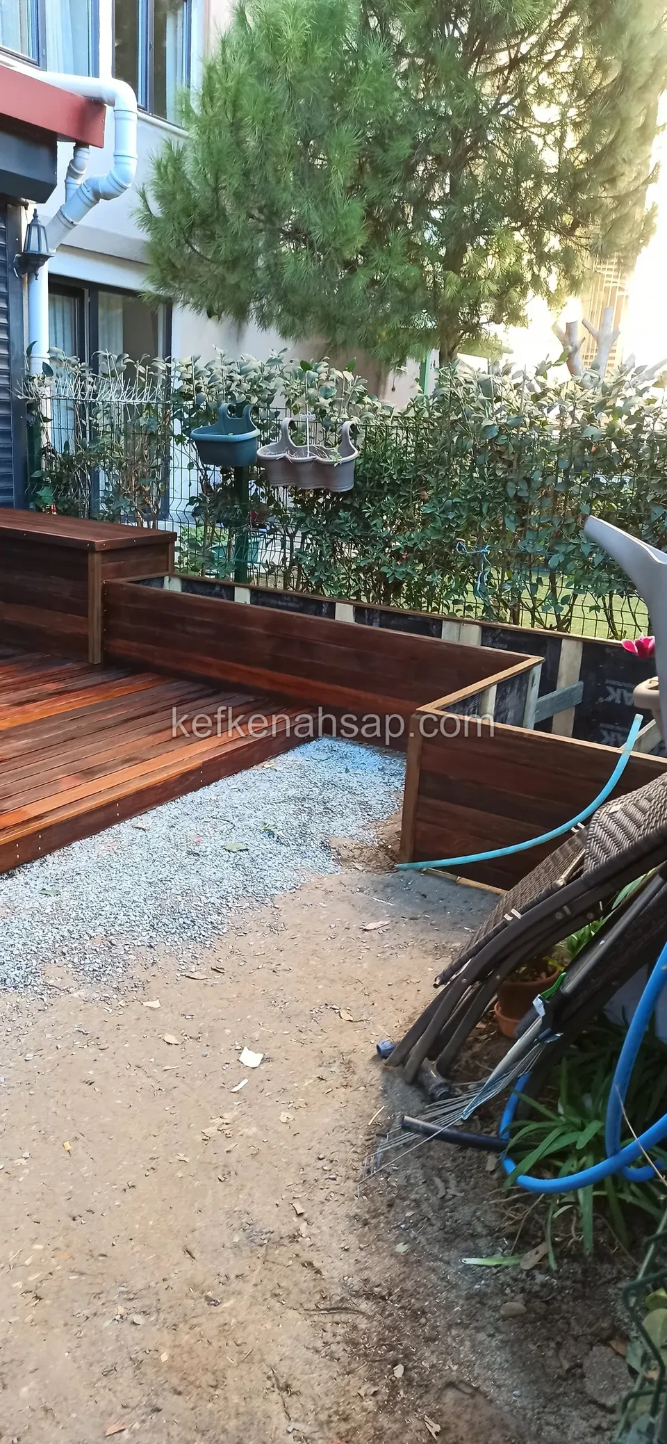 Dunkel rotbraunes Deck mit Holzpflanzkästen am Rand, rechts Schlauch und Pflanzen