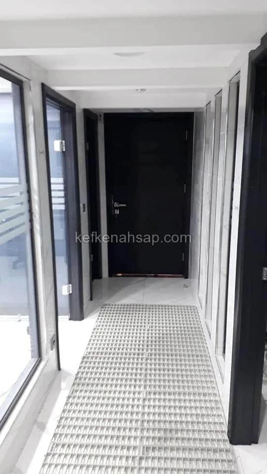 أبواب خشبية وإطارات مطلية بالأسود في ممر ضيق بأرضية بيضاء، وفي الوسط ممسحة شبكية