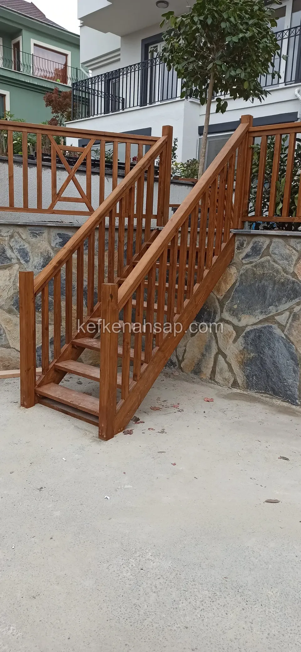 Taş kaplı duvarın önünde, dikey korkuluklu koyu kahve ahşap merdiven ve üstte aynı tonda korkuluk