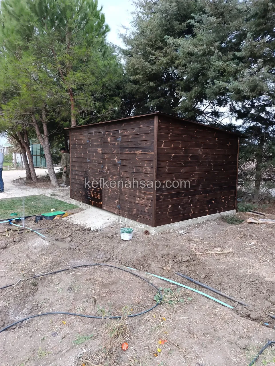Koyu vernikli ahşap kulübe; çevresinde serilmiş sulama hortumları ve kazılmış toprak, arkada çam ağaçları