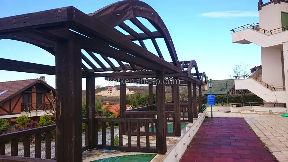 Kırmızı kaldırım kenarında koyu vernikli kemerli ahşap pergola ve çıtalı korkuluk, uzakta ahşap detaylı evler