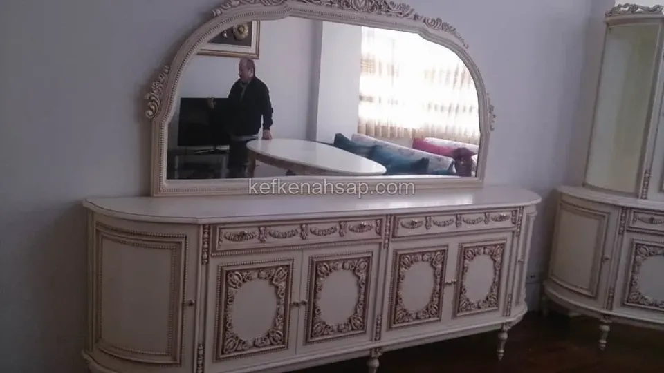 Krem boyalı, altın detaylı oymalı büfe; üzerinde kemerli aynası var, sağda aynı tarzda vitrin görünüyor.