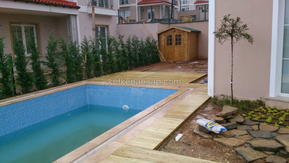 Heller Terrassenstreifen am Rand eines kleinen Pools, dahinter ein hölzernes Gartenhaus und eine Zypressenreihe