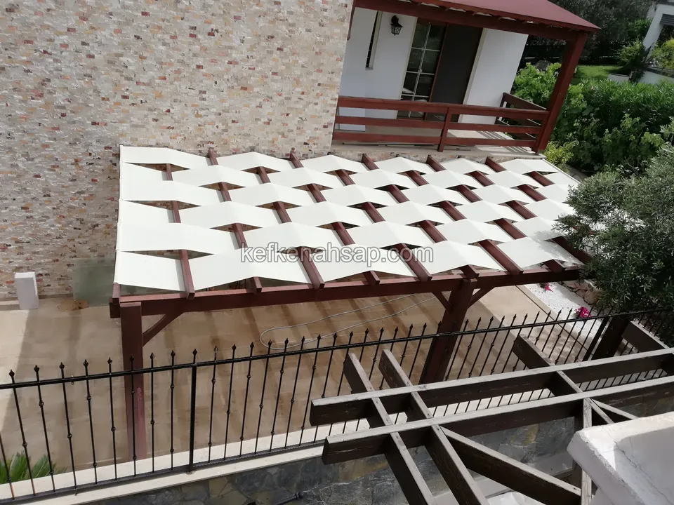Dark brown timber pergola in front of a stone-clad wall, with a cream fabric cover on top and an iron fence at the front