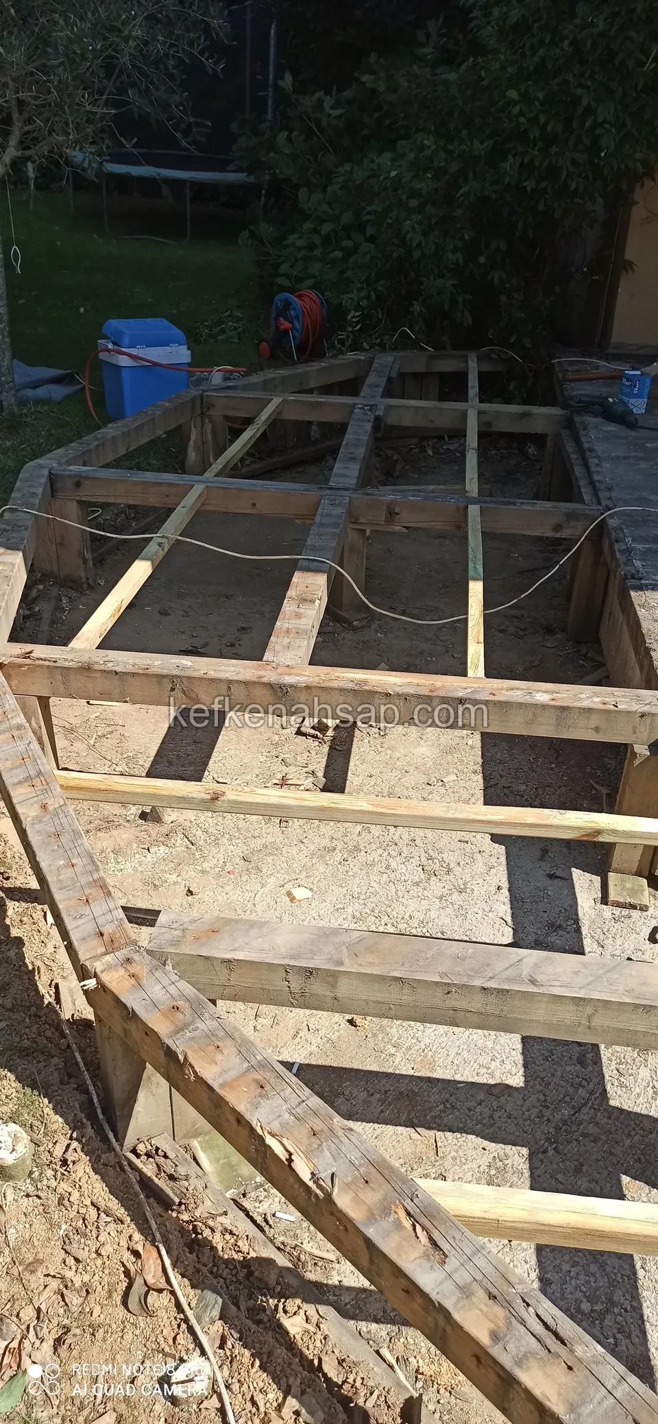 Kumlu zeminde, kalın ahşap kirişlerle kurulmuş deck alt çerçevesi; arkada bahçe ve mavi bir fıçı