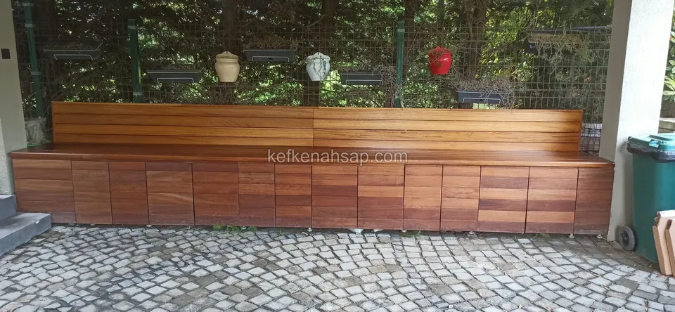 Küp taş zeminde yatay sırtlıklı uzun ahşap bank ve altında sıralı kapaklar; arkada saksılar asılı tel çit
