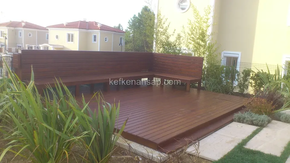 Arka duvar boyunca L şeklinde dönen ahşap bank ve önünde koyu kahverengi deck; ön tarafta bitkiler ve taş plakalar