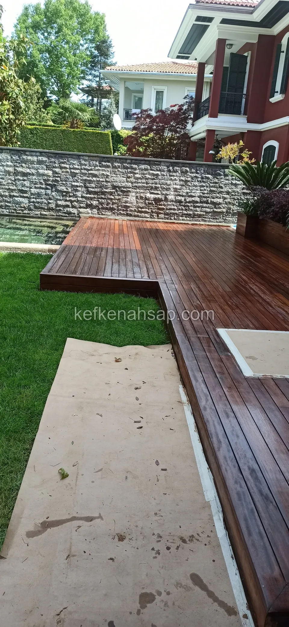 Dark brown polished deck turning in an L along the edge of a lawn, with a stone-clad wall behind