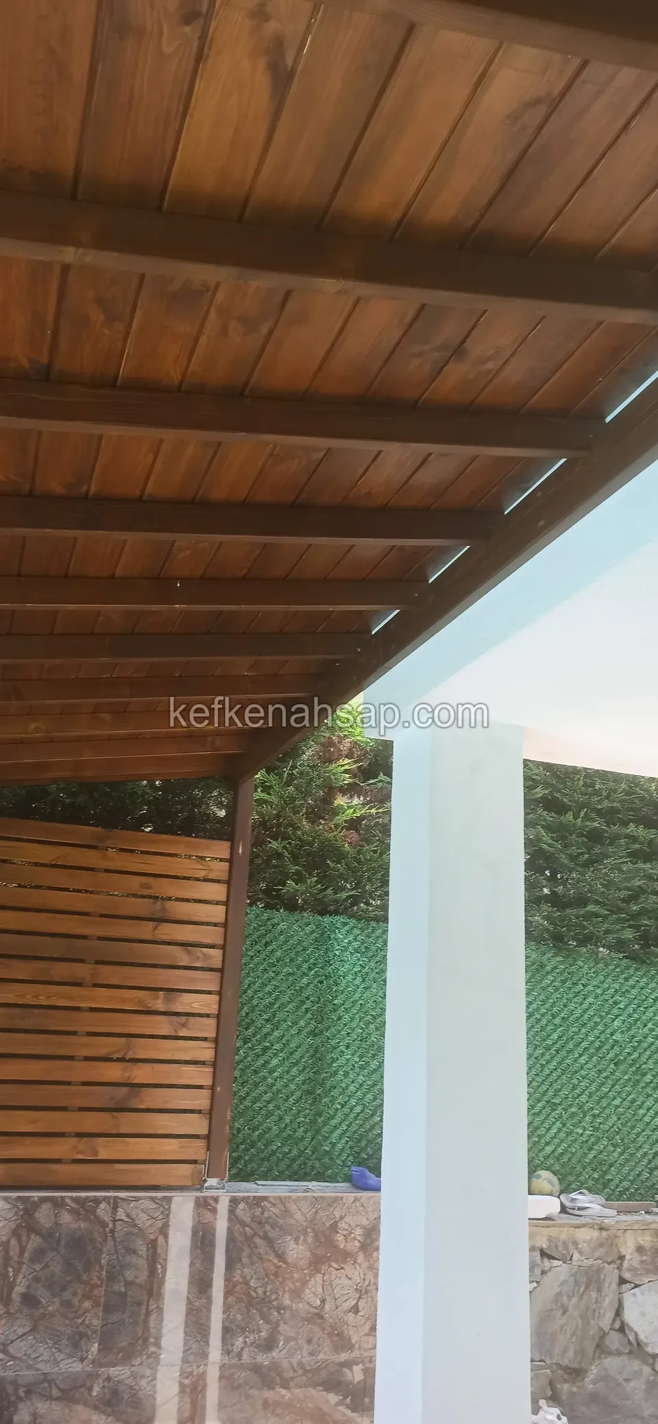 Çatının lambri kaplı alt yüzü, beyaz bir kolon ve yanındaki çıtalı ahşap panel, alttan görünüm