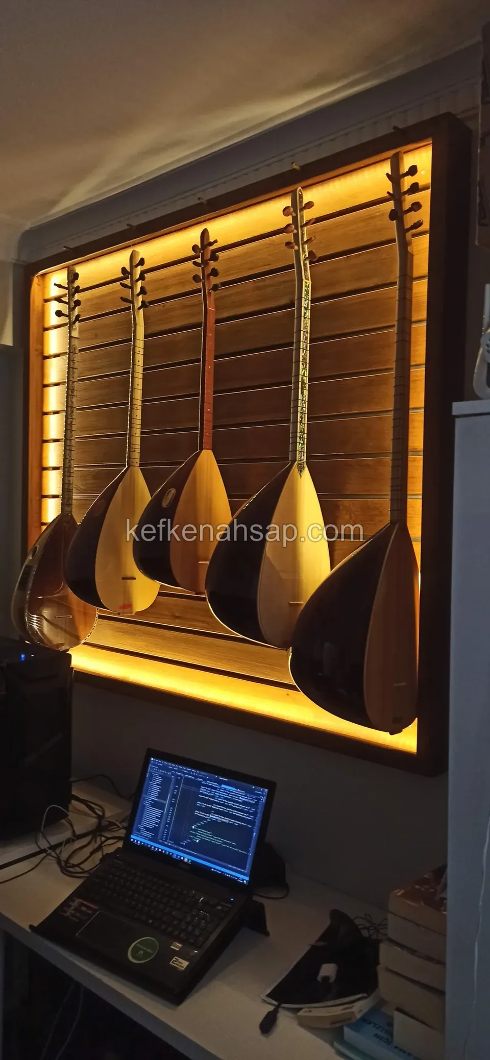 Duvarda koyu çerçeveli, arkadan sarı LED ile aydınlatılan ahşap panoda yan yana asılmış beş bağlama
