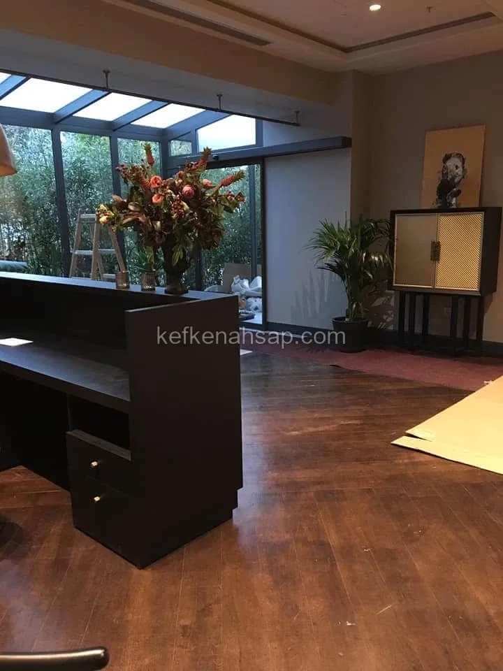 Cam tavanlı bir lobide koyu renk ahşap resepsiyon tezgâhı, üzerinde çiçek aranjmanı, arkada kapılı dolap ve parke zemin