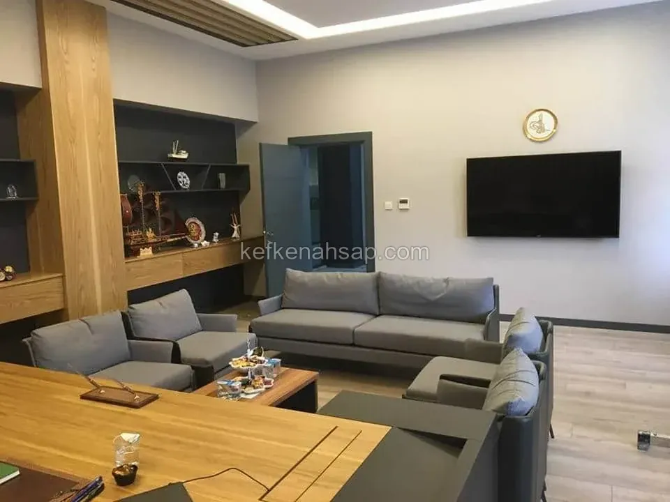 Açık meşe tonlu ahşap panel, gömme kitaplık ve çalışma masası olan ofiste gri koltuk takımı ile duvarda televizyon