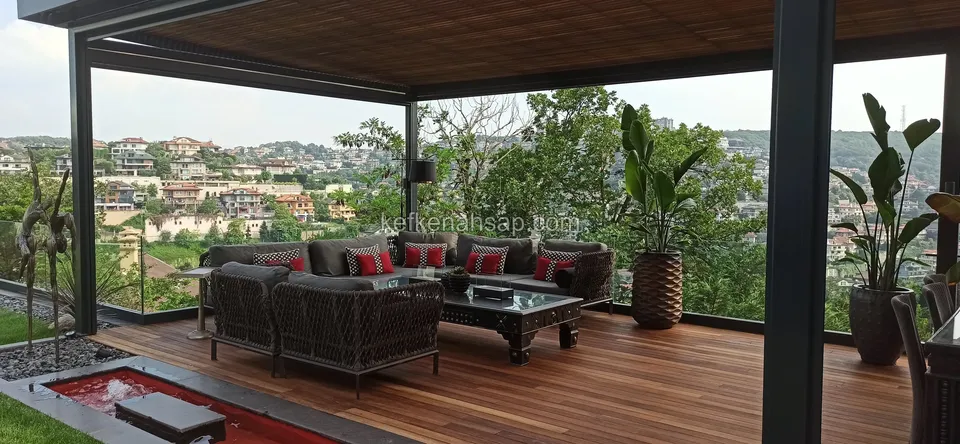Cam korkuluklu pergola altında, oturma grubu ve büyük saksılarla döşenmiş ahşap deck