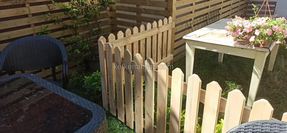 Heller Holzzaun aus spitzen Latten, der eine Terrassenecke vor einem Tisch mit Blumen und dunklen Rattansesseln einfasst