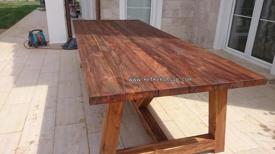 Long solid wood dining table with thick planks and sturdy braced legs on a travertine-paved terrace