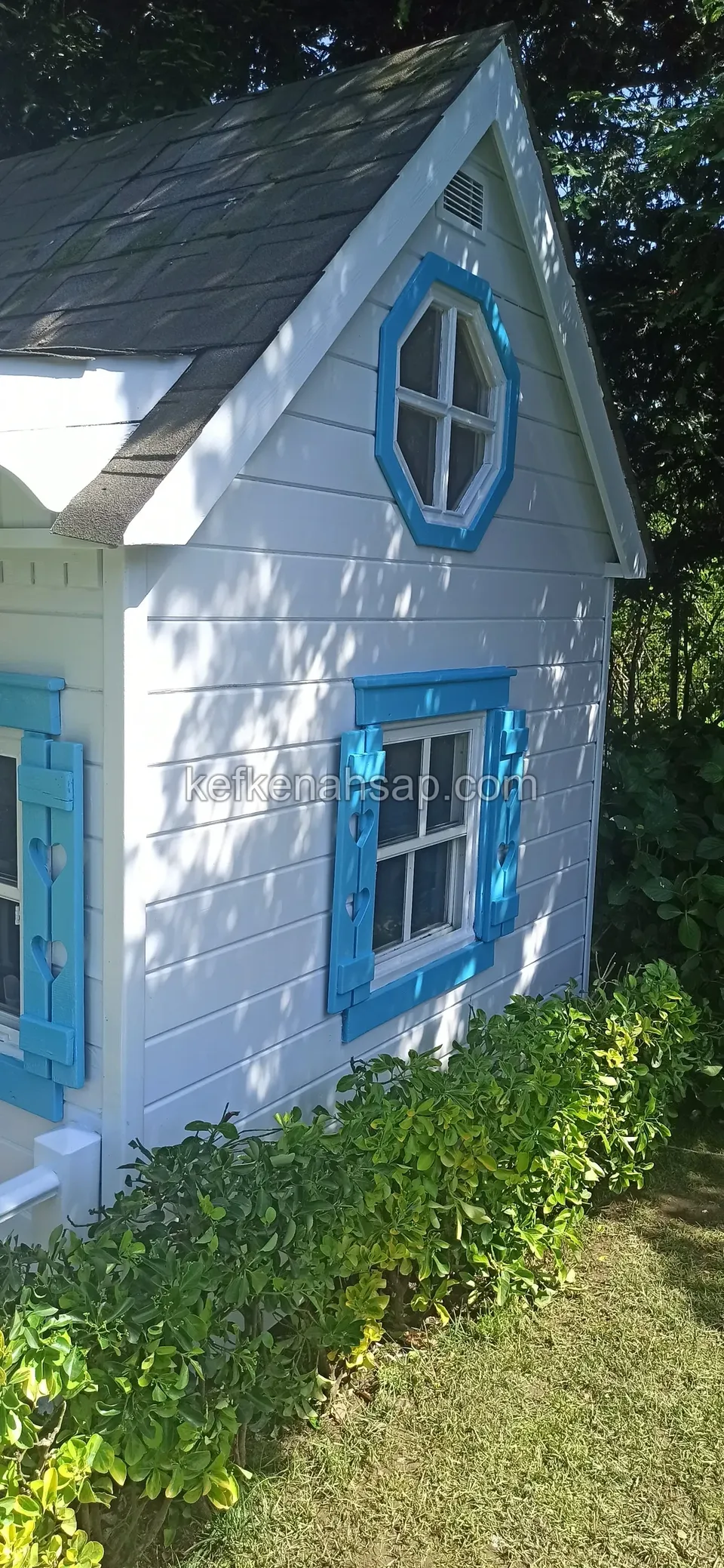 Beyaz boyalı, mavi çerçeveli sekizgen çatı penceresi ve kepenkli pencereleri olan ahşap oyun evi; yanında çalılık