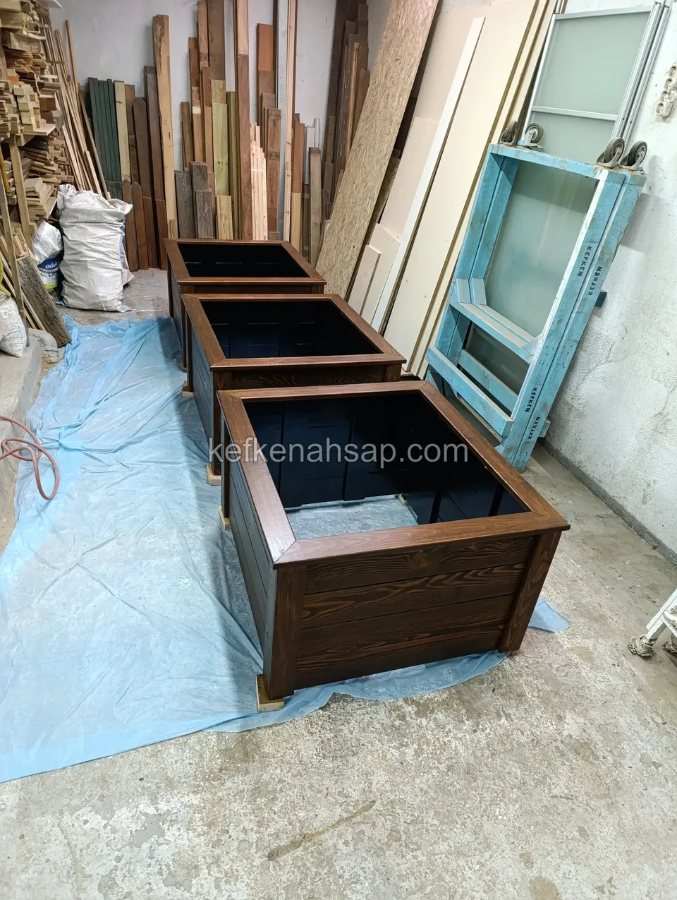 Three dark-varnished square timber planters lined up one behind another on blue plastic in the workshop