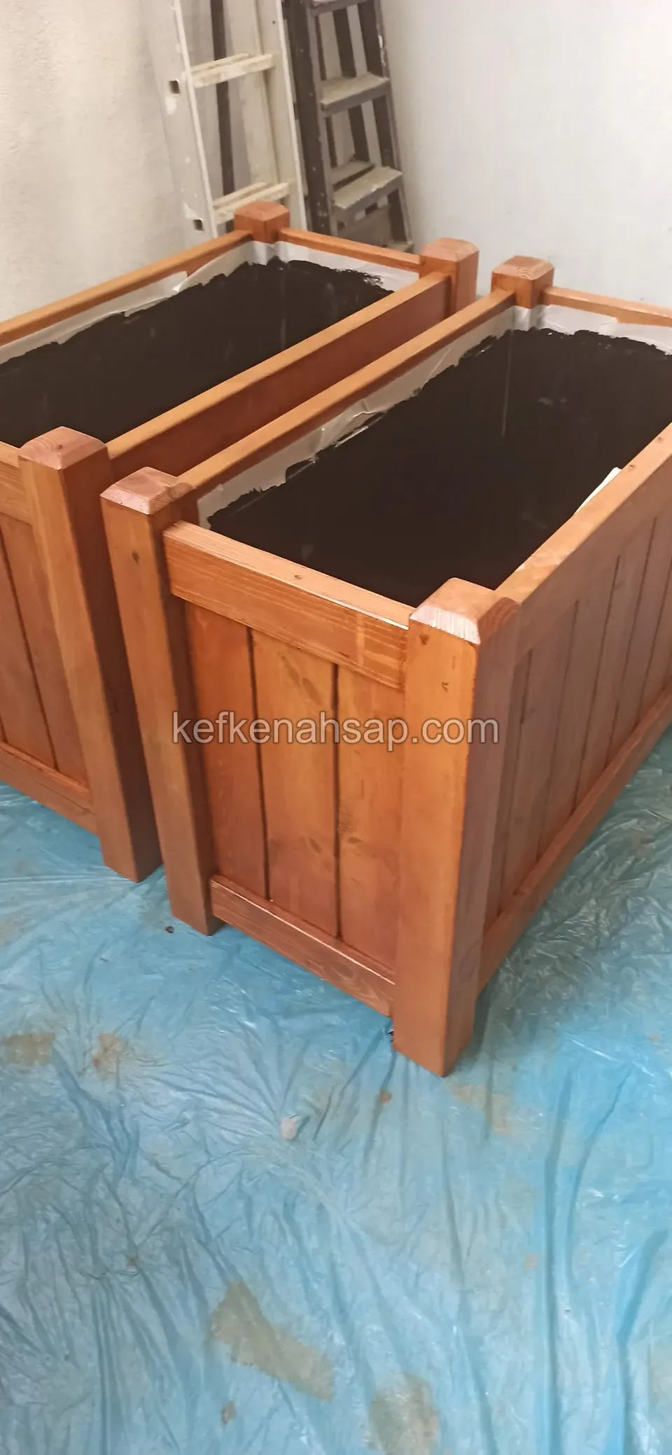 Two varnished vertical-board timber planters with black liners standing on blue plastic sheeting