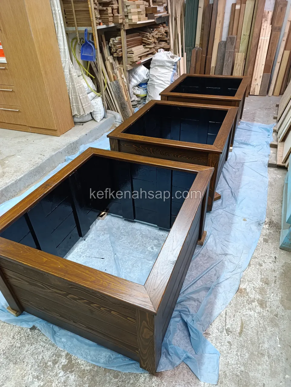 Three dark square timber planters on blue plastic, black-lined inside, with a timber rack behind