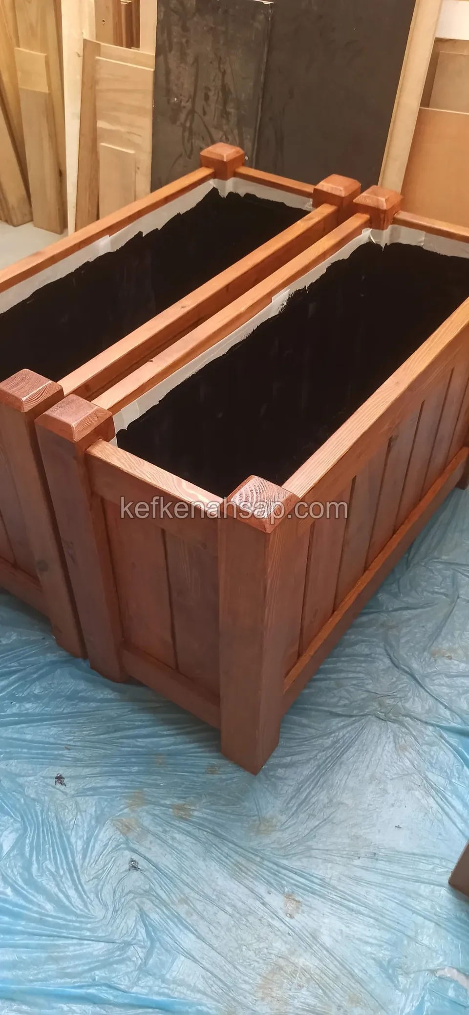 Two long varnished timber planters with black liners standing side by side on a blue sheet
