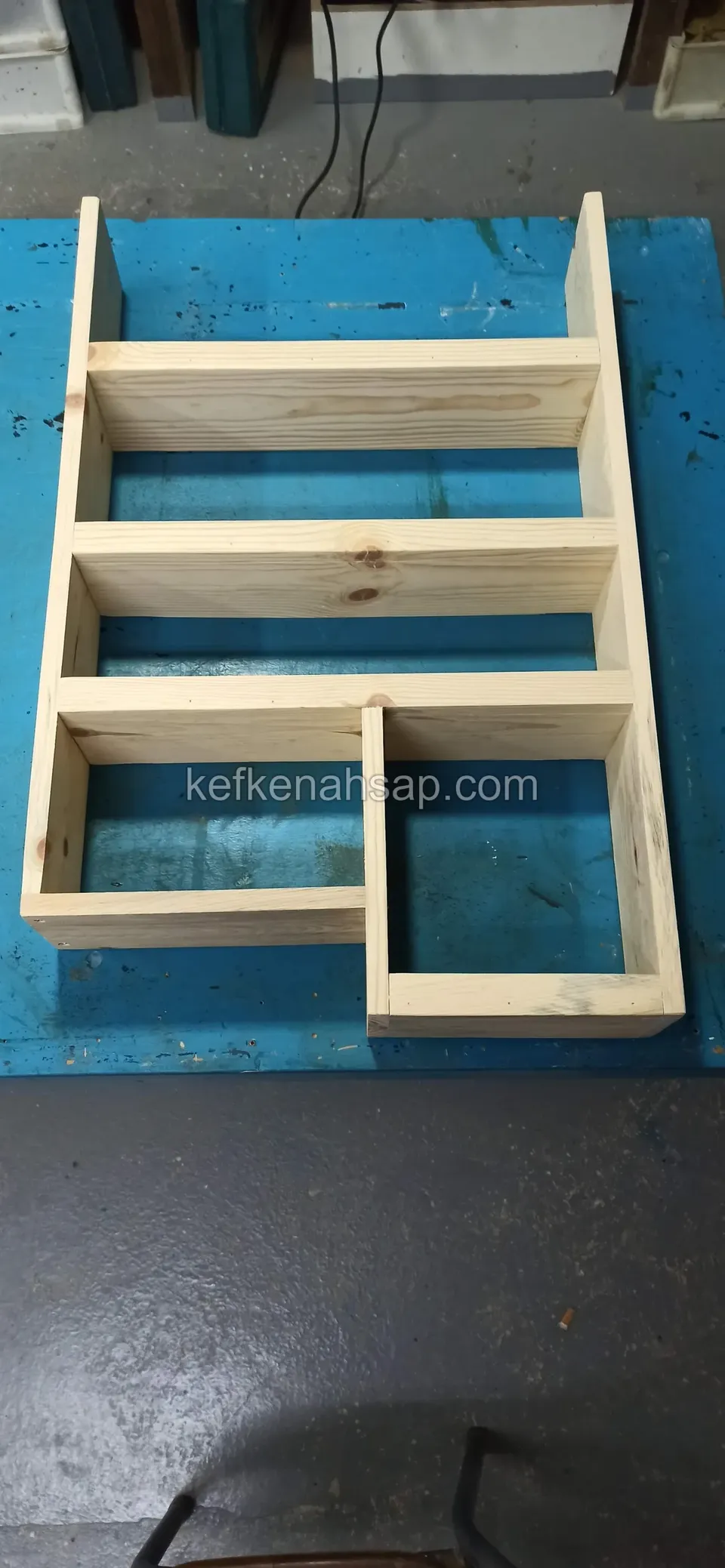 Ein helles Holzregalgerüst mit Fächern in Stufenform, aufrecht auf einer blauen Werkbank stehend