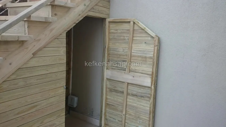 Merdiven altında yatay tahta kaplama ve açık duran panjurlu ahşap kapak; içeride bir pano görünüyor