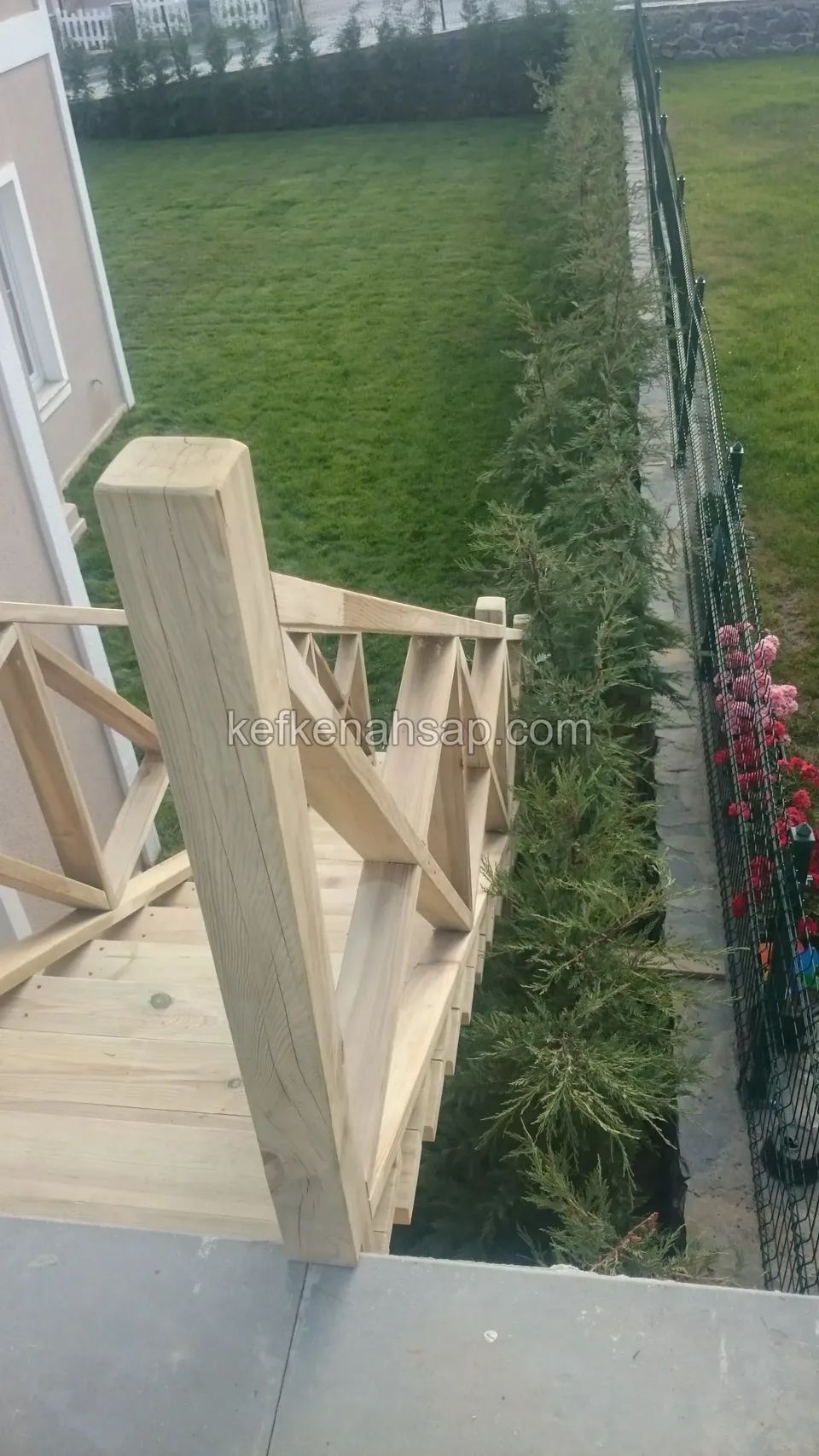 Çimlik kenarındaki ahşap merdivenin başında kalın bir direk ve X çaprazlı açık tonlu korkuluk, yanda servi sırası ve tel çit