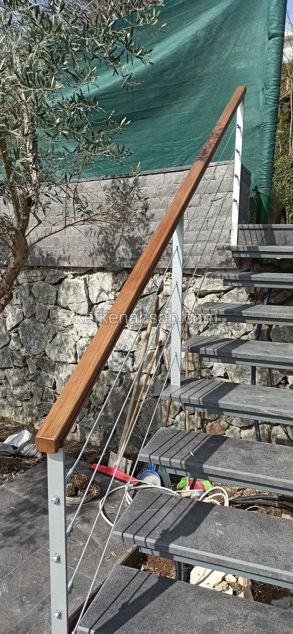 Metal basamaklı merdivende ahşap küpeşte ve ince halatlı korkuluk; yanda taş duvar ve zeytin ağacı