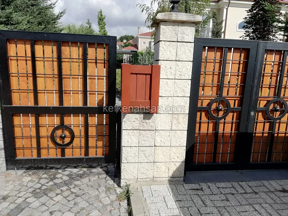 Siyah çerçeveli, ahşap dolgulu kapının daire motifli kanatları ve beyaz taş sütunun üstündeki ahşap posta kutusu