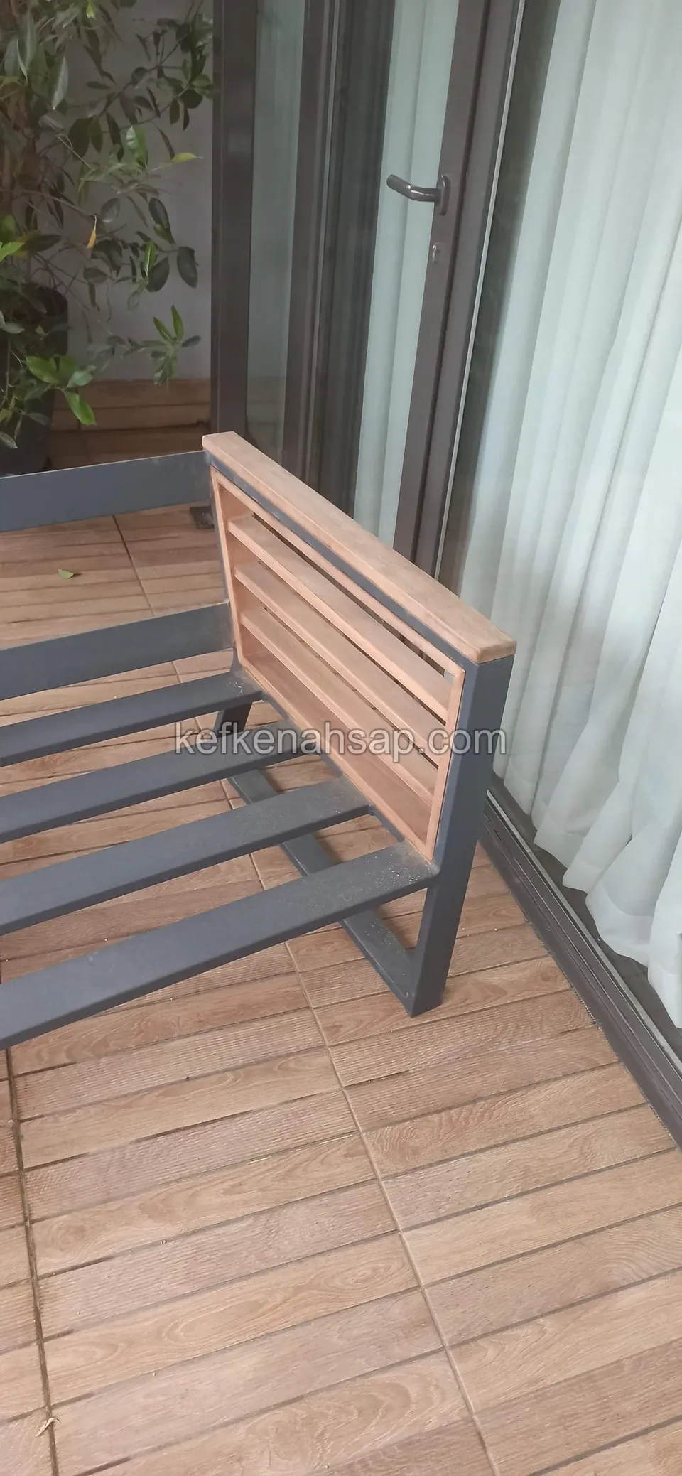 Cam kapılı bir terasta, gri metal çerçeveye çıtalı ahşap sırt ve oturak takılmış, minderi olmayan bir koltuk duruyor