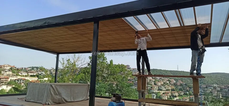 Two craftsmen on a platform fitting timber ceiling slats and a clear panel to a metal-framed pergola, with houses behind